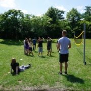 Mehrere Jugendliche stehen im Park vor einem Volleyballnetz in einem Kreis. Ein Jugendlicher im weißen Tshirt und ein Jugendlicher im schwarzen Tshirt stehen bzw. sitzen wartend vor den anderen Jugendlichen